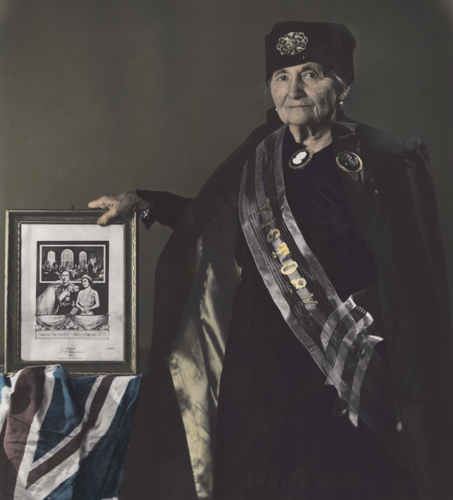 Photographie colorisée de Roza Brown en femme âgée portant une cape, une robe et un chapeau sombres. Une ceinture portant le mot Victoire en lettres dorées est drapée sur le devant de sa robe. Elle regarde la caméra en tenant une image encadrée du roi George VI et de son épouse la reine Elizabeth. Une table à sa gauche est drapée d’un drapeau britannique.