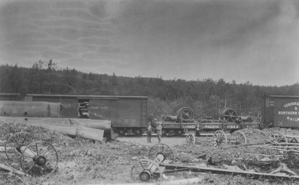 Photographie en noir et blanc de grandes pièces de machines de mines sur un wagon de chemin de fer. Deux hommes se tiennent devant le wagon. Au premier plan, on aperçoit des pièces de métal, de grands wagons et des roues.