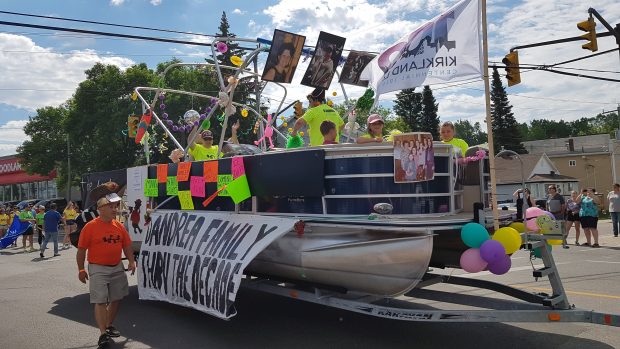 Photographie couleur d’un grand bateau ponton sur une remorque tirée dans la rue comme un char allégorique dans un défilé. Le char allégorique est décoré d’une bannière intitulée « La famille D’Andrea à travers les décennies » accrochée sur le côté avec d’autres décorations. Au moins cinq personnes sont visibles sur le char allégorique.