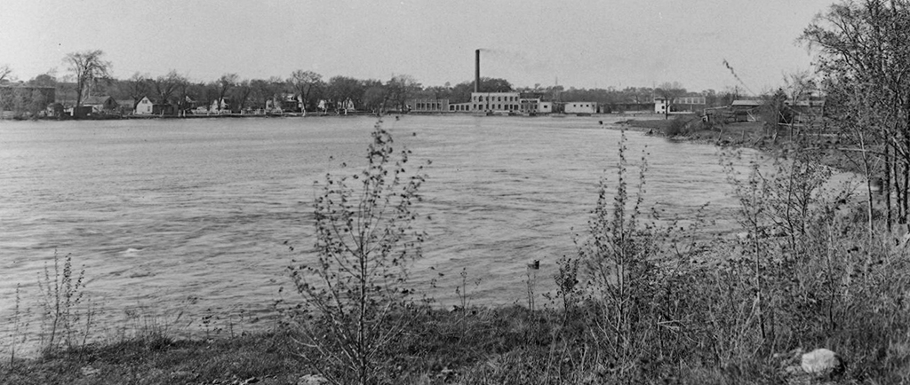 Photographie noir et blanc prise près du bassin d’eau derrière l’église de la Visitation. À droite sur la digue se trouve l’usine de la Back River Power Company avec sa grande cheminée. À gauche, les maisons de la rue de l’Île-de-la-Visitation.