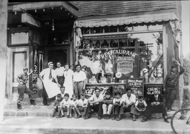 Plusieurs garçons avec quelques hommes sont assis devant la vitrine du restaurant de Jeff Geoffrion. Debout à leur gauche, quatre hommes et une femme semblent composer le personnel du commerce. Il y a une marquise éclairée devant la porte d’entrée.