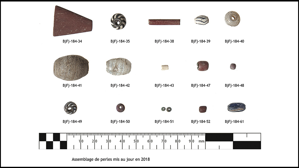 Assemblage de perles retrouvées par des archéologues lors de fouilles en 2018. Il est constitué de treize perles en verre et deux perles façonnées en argilite rouge. Au bas du tableau, une échelle en millimètres.