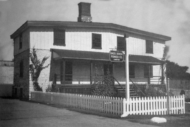 Derrière une clôture en planches de bois peintes en blanc, le bâtiment qui abrite les bureaux de la Back Power Company en 1942. La maçonnerie est visible au rez-de-chaussée, mais un revêtement mural différent est présent à l’étage. Une galerie avec une toiture inclinée a été ajoutée.