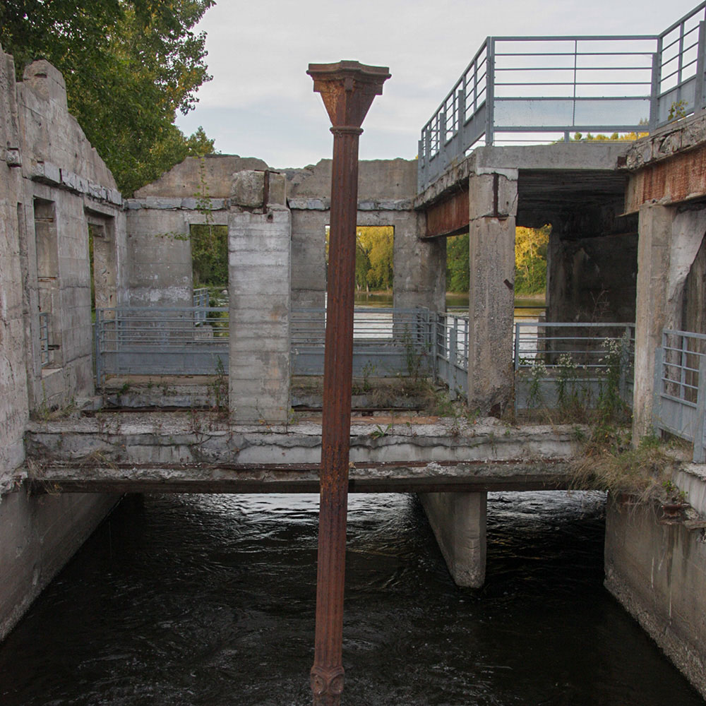 Une colonne de fer demeure plantée dans l’eau isolée parmi les vestiges des bâtiments industriels.