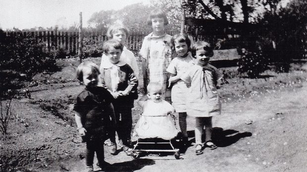 Six enfants de la famille Danis entourent un plus jeune qui est dans une marchette. Ils sont dans un jardin fermé par une clôture de bois.