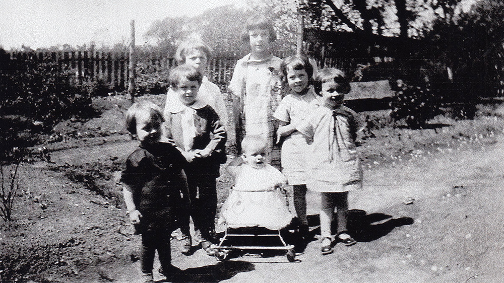 Six enfants de la famille Danis entourent un plus jeune qui est dans une marchette. Ils sont dans un jardin fermé par une clôture de bois.