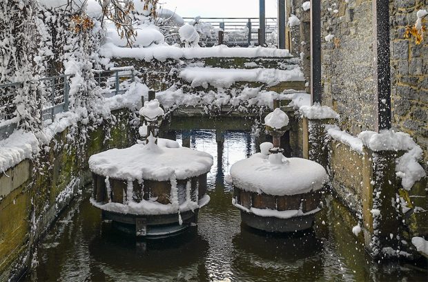 Après une chute de neige, deux turbines hydrauliques sont recouvertes de blanc. Elles sont aujourd’hui immobiles au-dessus du niveau de l’eau qui passe à travers la digue. À leur droite, un ancien mur de maçonnerie.