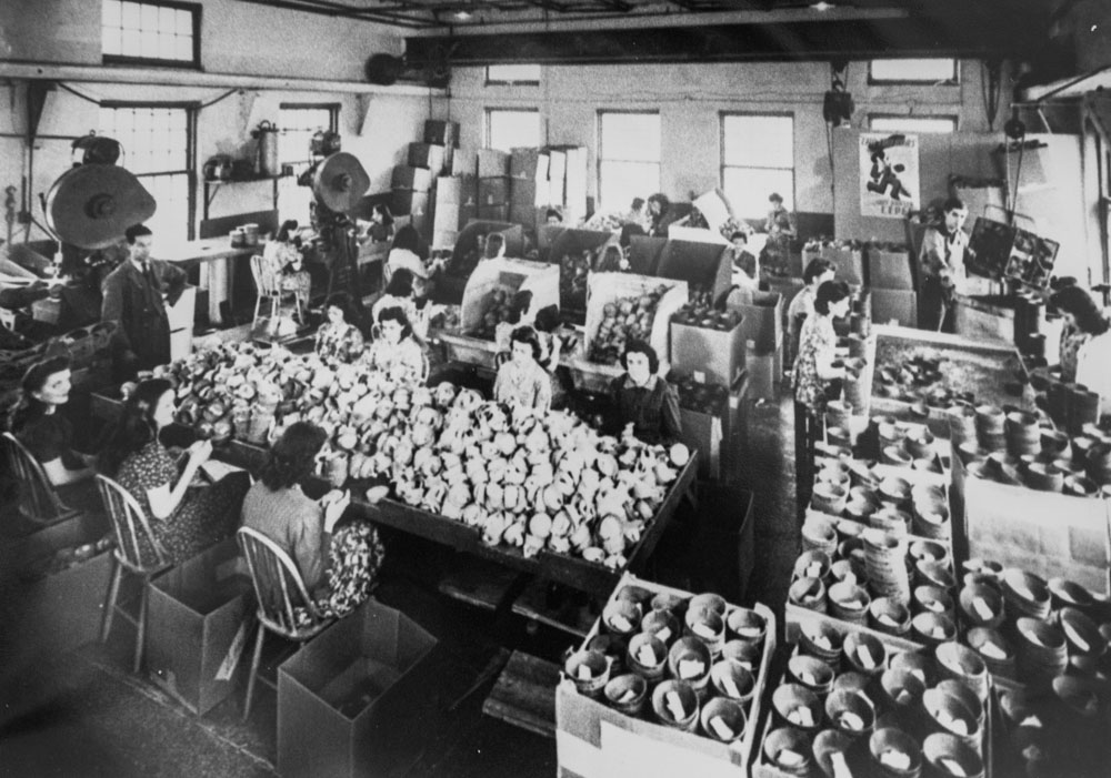 À l'étage de l'usine, des femmes préparent des emballages de munitions, 1942.Vue intérieure d’un atelier de la Back River Power Company en 1942. Des ouvrières assises autour de grandes tables préparent des emballages cartonnés cylindriques pour les munitions qui seront envoyées sur les fronts européens. Debout à la gauche, un homme observe leur travail. À la droite, des boites pleines de cylindres.