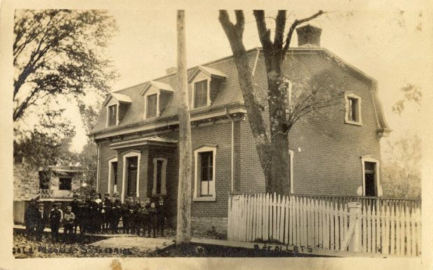 En 1900, l’école du village, au centre, ressemble à une grande maison en briques avec un toit mansardé et des fenêtres à lucarnes. À sa gauche, un groupe de garçons avec leur enseignant. À sa droite, une clôture de bois.