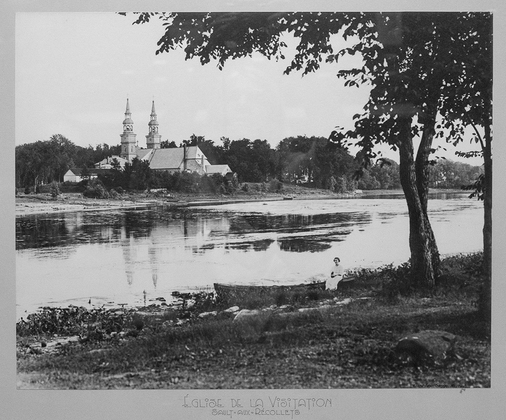 Une dame vêtue de blanc est assise sur une chaloupe sous un arbre au bord du bassin de la digue. À l’arrière-plan, l’église de la Visitation avec ses deux clochers sur la rive de l’île de Montréal. Le village avec ses nombreux arbres a conservé un air champêtre.
