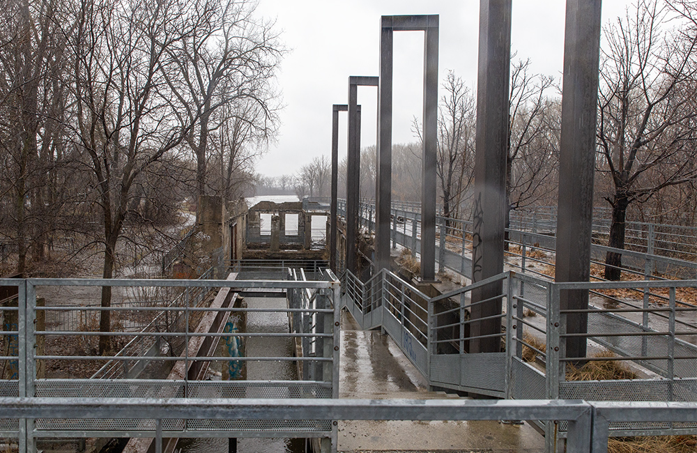 Vue prise dans l’axe du belvédère sur la rivière à partir de la rue du Pont, par une journée pluvieuse de printemps. À gauche, des arbres dénudés, à droite, les structures d’acier qui rappellent le volume des anciennes constructions. Au fond, l’eau de la rivière.