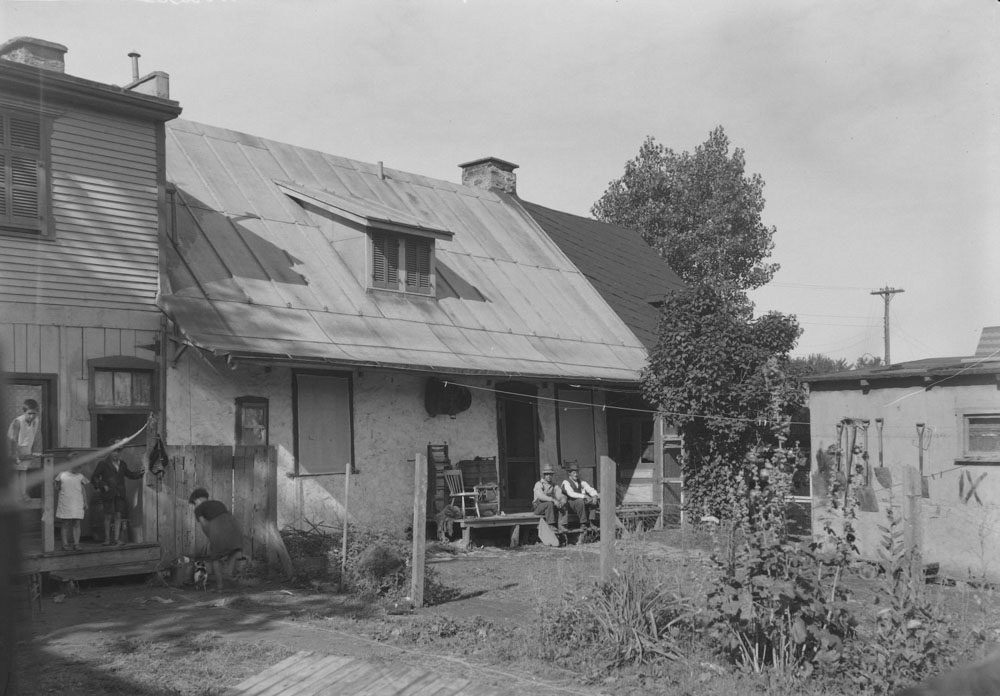 À gauche, des enfants jouent dans la cour arrière du logement au 10861, rue du Pressoir. C’est une construction à toit plat à deux étages qui s’est ajoutée au bâtiment du Pressoir. Au centre, deux adultes causent sur le balcon du 10865. Le Pressoir est alors divisé en deux logements avec des toitures à double pente de couleurs différentes.