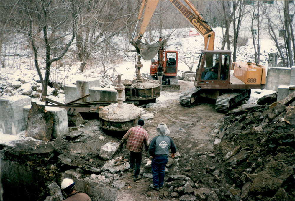 Au centre, deux hommes regardent l’opérateur d’une excavatrice sur chenille retirer des turbines de leur emplacement par une journée enneigée. Le bassin a été asséché pour permettre le réaménagement de la digue des moulins à la fin des années 1990.