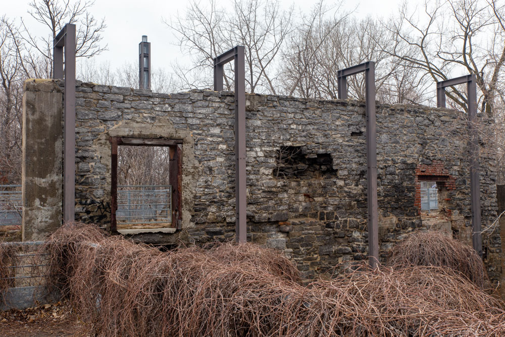 Sur la digue des moulins, un ancien mur restauré en maçonnerie est cerclé de quatre structures d’acier. Elles représentent le volume des anciens bâtiments industriels. Derrière le mur, la tour d’acier qui occupe l’emplacement d’une haute cheminée d’usine en briques disparue.