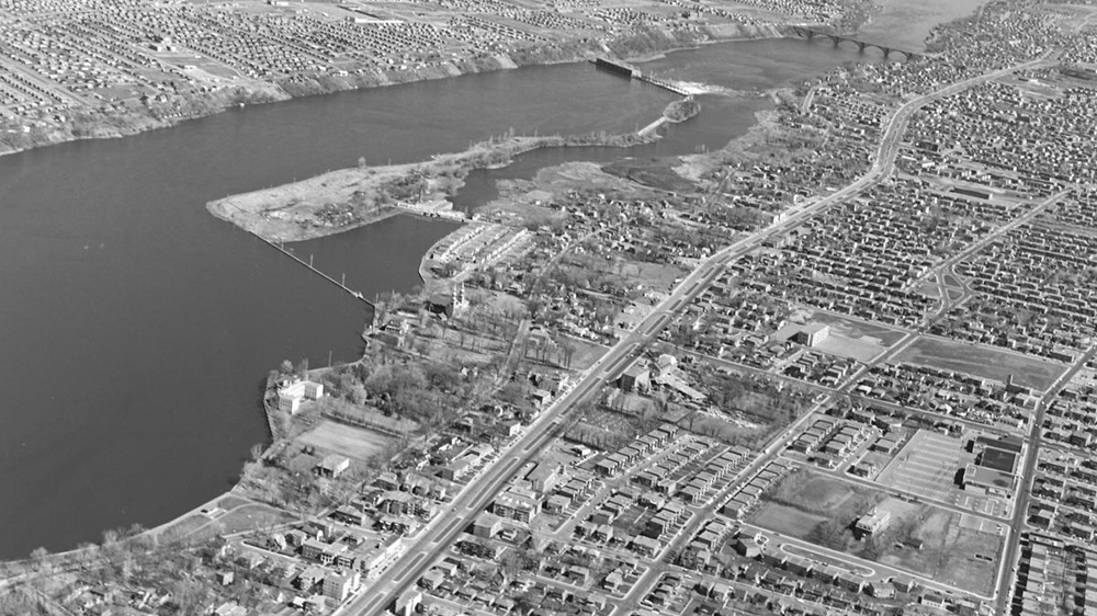 Vue aérienne oblique de la rivière des Prairies en 1963. À l’avant-plan, les nouveaux quartiers résidentiels denses du Sault-au-Récollet. Dans la rivière, l’île de la Visitation, la centrale hydroélectrique et, en haut à droite, le pont Pie-IX à Montréal-Nord.