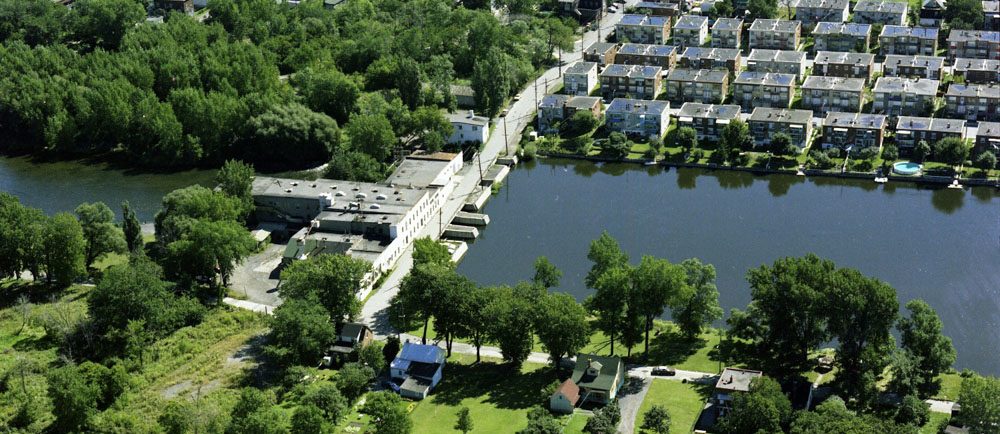 Vue aérienne du site des Moulins en 1976. Au centre: l’eau de la rivière des Prairies traverse la digue et ses bâtiments industriels désaffectés. À l’avant-plan et à gauche, le territoire du futur parc-nature de l’île de l’Île-de-la-Visitation. À droite, des duplex en rangée.
