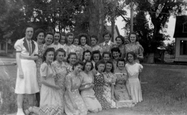 Vingt-cinq ouvrières, réparties en trois rangées, posent sur la rue de l’Île-de-la-Visitation en 1941. Elles sont vêtues de robes différentes. Derrière elles, des arbres et des maisons de l’île.