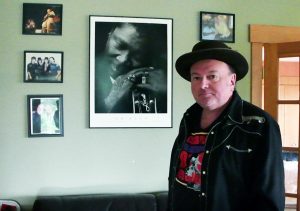 Photo en couleur d'un homme vêtu d'une veste noire sur un t-shirt coloré, portant un chapeau à bords noirs, debout devant un mur clair décoré de photos encadrées de personnes, certaines tenant des instruments de musique.