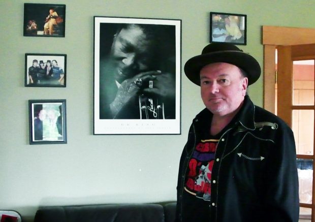Photo en couleur d'un homme vêtu d'une veste noire sur un t-shirt coloré, portant un chapeau à bords noirs, debout devant un mur clair décoré de photos encadrées de personnes, certaines tenant des instruments de musique.