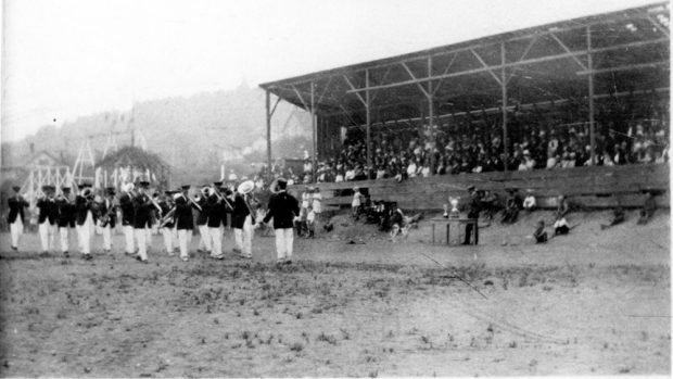 Photo en noir et blanc de membres d'un groupe de musique jouant à l'extérieur, sous le regard de spectateurs dans la tribune.