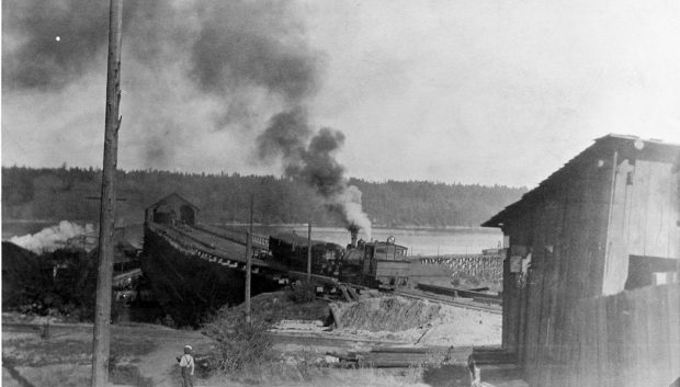Photo en noir et blanc d'installations industrielles charbonnières situées sur le front de mer. Un jeune garçon observe une machine à vapeur manœuvrant des wagons de charbon chargés sur une voie ferrée surélevée. En arrière-plan, les eaux d'un port et un grand quai sont visibles.
