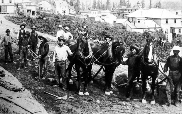 Photo en noir et blanc représentant un groupe de 12 hommes, dont deux sont asiatiques et les autres caucasiens, ainsi que trois chevaux attelés, debout sur une route de terre partiellement nivelée.