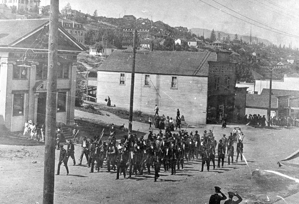 Photo en noir et blanc d'une rue non pavée de la ville, le long de laquelle marchent des soldats armés encadrant un groupe de civils.