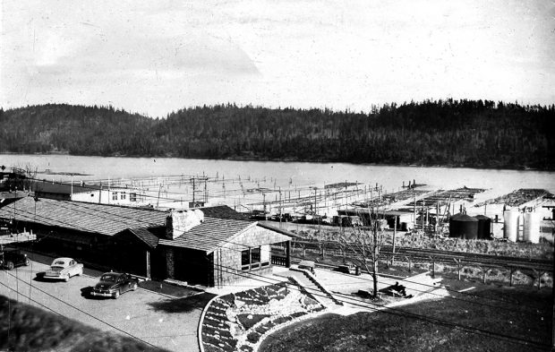 Photographie en noir et blanc d'un immeuble de bureaux à un étage avec plusieurs voitures garées devant. Des barrages de troncs sont ordonnés et flottent dans les eaux du port en arrière-plan.