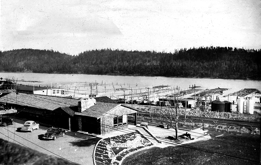 Photographie en noir et blanc d'un immeuble de bureaux à un étage avec plusieurs voitures garées devant. Des barrages de troncs sont ordonnés et flottent dans les eaux du port en arrière-plan.