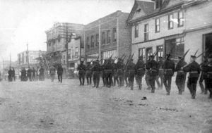 Photo en noir et blanc de soldats, fusils sur l'épaule, marchant le long d'une rue non pavée de la ville.