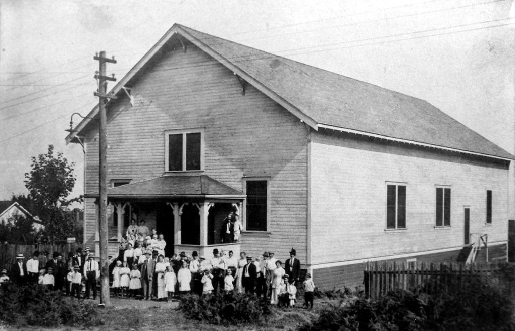 Photo en noir et blanc d'un bâtiment rectangulaire en bois à un étage avec un toit à pignon et un porche à l'avant, autour duquel un groupe d'hommes, de femmes et d'enfants bien vêtus se tiennent face au photographe.