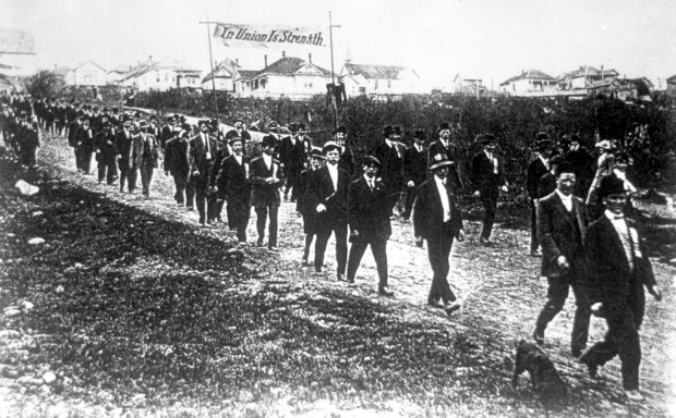 Photo en noir et blanc d'hommes en vêtements civils défilant en descente sur une rue non pavée, portant une bannière avec l'inscription « L'union fait la force ». Certains hommes portent des épinglettes bien visibles.