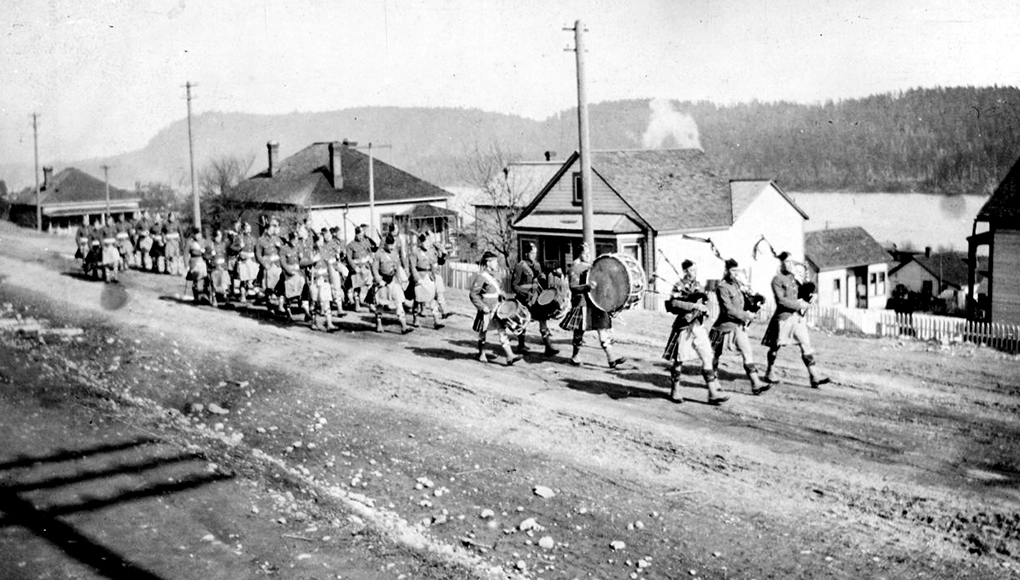 Photo en noir et blanc d'une formation de soldats en kilt, dirigée par une fanfare composée de trois joueurs de cornemuse et de trois tambours, marchant le long d'une rue non pavée bordée de maisons en bois détachées, entre lesquelles on aperçoit les eaux d'un port.
