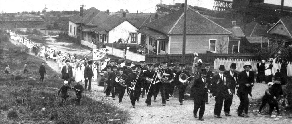  Photographie en noir et blanc d'une rue en contrebas, remontée par une fanfare menant un cortège de femmes et d'enfants. Les eaux d'un port forment l'arrière-plan.