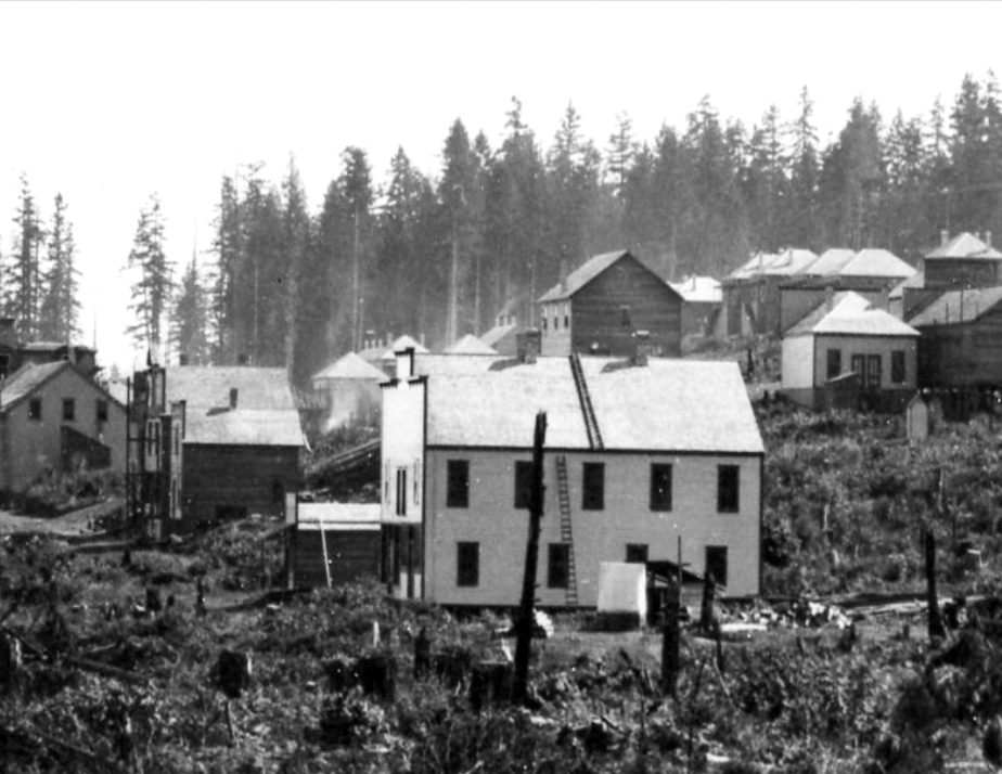 Photo en noir et blanc d'un bâtiment rectangulaire en bois à deux étages, de couleur claire, avec un toit en pente et de grandes fenêtres rectangulaires disposées de manière décalée entre les niveaux, entouré d'un espace ouvert occupé par des buissons et des souches d'arbres. D'autres bâtiments en bois de construction similaire se trouvent à l'arrière-plan.