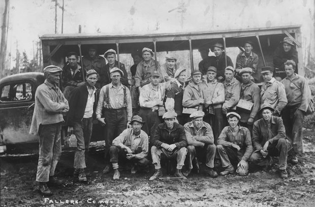 Photographie en noir et blanc d’un groupe de 26 hommes en vêtements de travail, certains portant des seaux à lunch, posant devant un camion transformé en autobus rudimentaire à parois ouvertes.