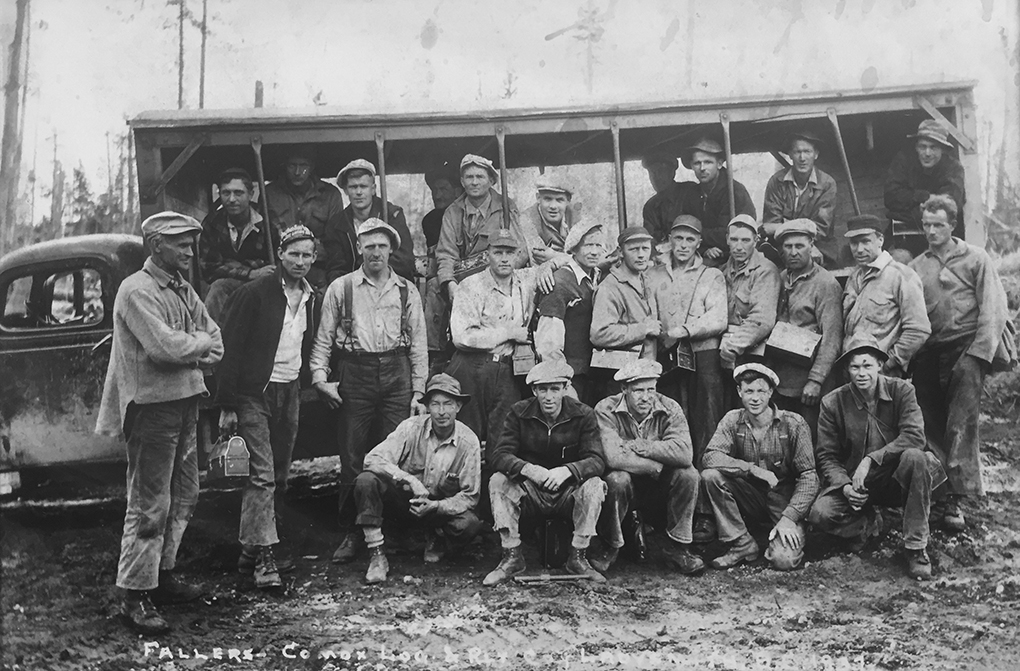 Photographie en noir et blanc d’un groupe de 26 hommes en vêtements de travail, certains portant des seaux à lunch, posant devant un camion transformé en autobus rudimentaire à parois ouvertes.