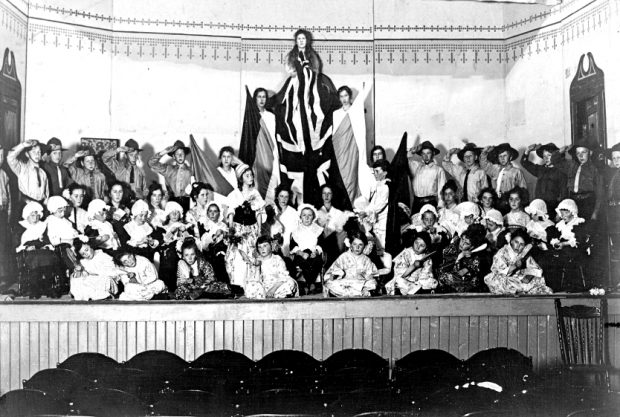 Photo en noir et blanc d'un grand groupe d'enfants acteurs costumés, disposés sur scène en trois rangées : à genoux devant, assis au milieu et debout à l'arrière. Au dernier rang, trois actrices surélevées tiennent des drapeaux verticaux, avec un Union Jack au centre, surplombant l'ensemble.