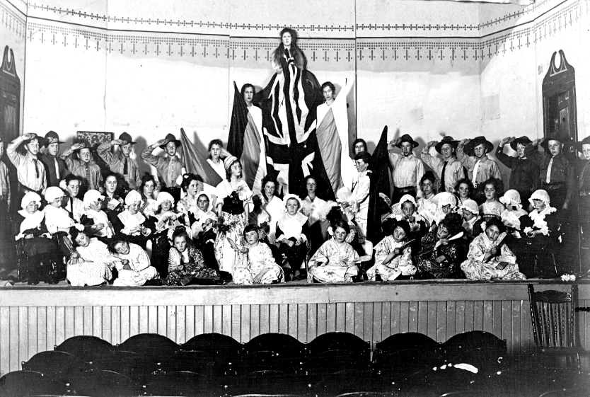 Photo en noir et blanc d'un grand groupe d'enfants acteurs costumés, disposés sur scène en trois rangées : à genoux devant, assis au milieu et debout à l'arrière. Au dernier rang, trois actrices surélevées tiennent des drapeaux verticaux, avec un Union Jack au centre, surplombant l'ensemble.