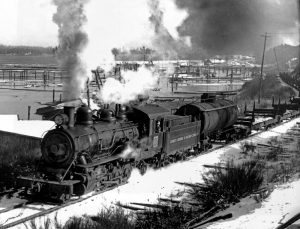 Photographie en noir et blanc d'une locomotive à vapeur, de la fumée et de la vapeur s'échappant de sa cheminée et de la vapeur s'échappant de son sifflet, tirant une file de wagons plats vides. En arrière-plan, les eaux du port de Ladysmith, avec des pieux enfoncés délimitant les zones de bouillage.