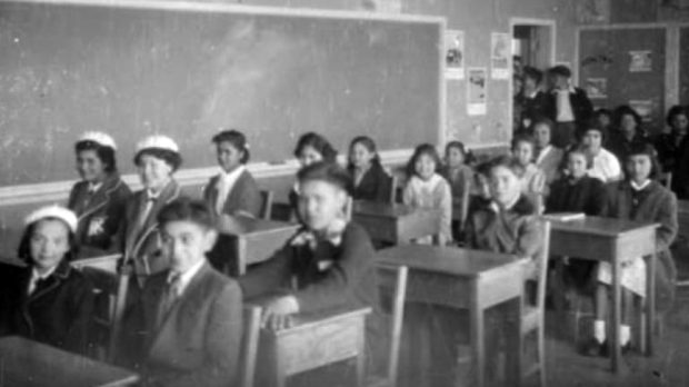 Photo en noir et blanc montrant des élèves vêtus de façon soignée assis à leur bureau dans une salle de classe.