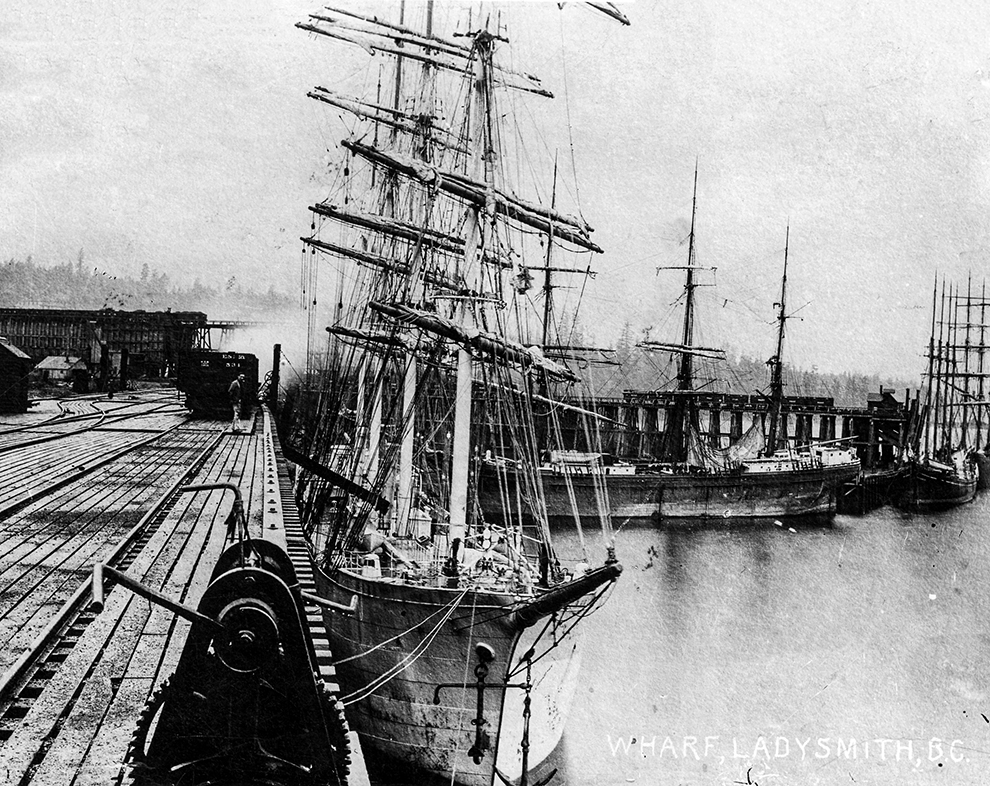 Photo en noir et blanc de quatre navires à voile océaniques, voiles repliées, amarrés le long de deux grands quais en bois.