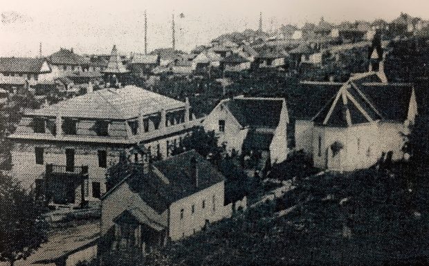 Vue panoramique en noir et blanc d'une ville, avec un groupe de quatre bâtiments au premier plan, dont l'un présente une architecture d'église typique.