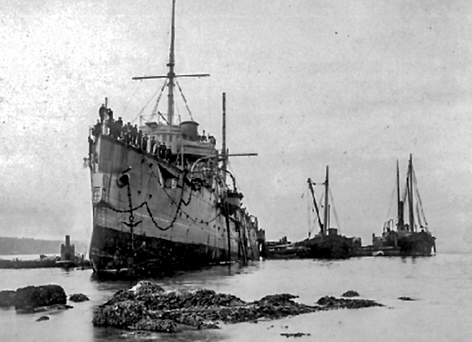Photo en noir et blanc montrant une vue oblique du côté bâbord à la ligne de flottaison du bow d'un cuirassé à coque en fer échoué sur des rochers. On voit des membres d'équipage le long des rambardes du pont. Plusieurs petits navires sont présents pour porter assistance.