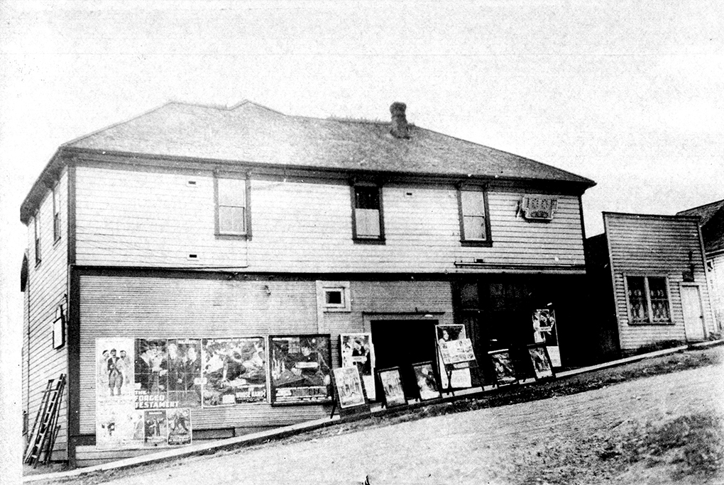 Photo en noir et blanc d'un bâtiment en bois à deux étages vu de l'autre côté d'une rue en pente raide non pavée. De nombreux panneaux publicitaires ornent les murs au niveau de la rue.