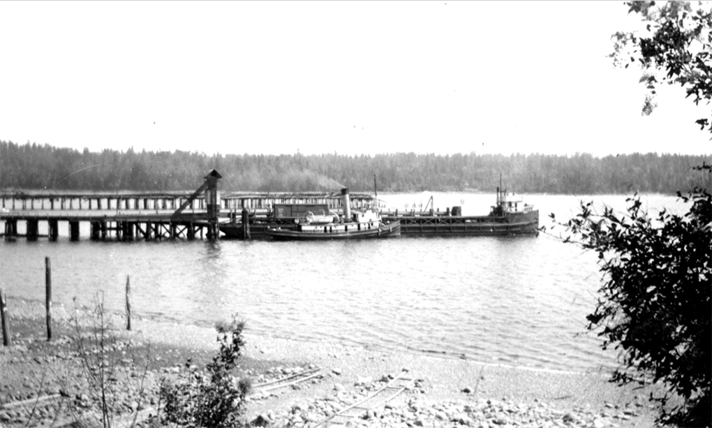 Photographie en noir et blanc d'un remorqueur à cheminée unique approchant une barge partiellement chargée à côté d'un quai.