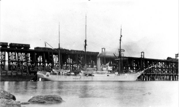 Photo en noir et blanc d'un navire à trois mâts, voiles repliées, avec une cheminée bien visible et fumante au milieu du navire, amarré à côté et écrasé par les pilotis d'un grand quai en bois, sur lequel se trouvent des wagons de charbon en bois, chargés et vides.