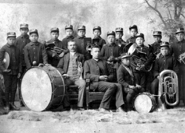 Photo en noir et blanc de 15 garçons des Premières Nations, en uniforme simple avec une casquette, portant des instruments de fanfare, debout derrière deux hommes caucasiens assis, dont l'un est un prêtre.