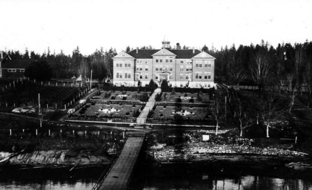 Photo aérienne en noir et blanc d'un quai en bois menant du rivage à un chemin en escalier traversant des jardins soignés et symétriques, devant un imposant bâtiment en brique de trois étages.