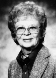 Portrait en noir et blanc, vue de la tête et des épaules, d'une femme d'âge moyen souriante, aux cheveux bouclés et portant des lunettes, vêtue d'une veste en laine sur une chemise foncée à boutons et d'un foulard autour du cou.